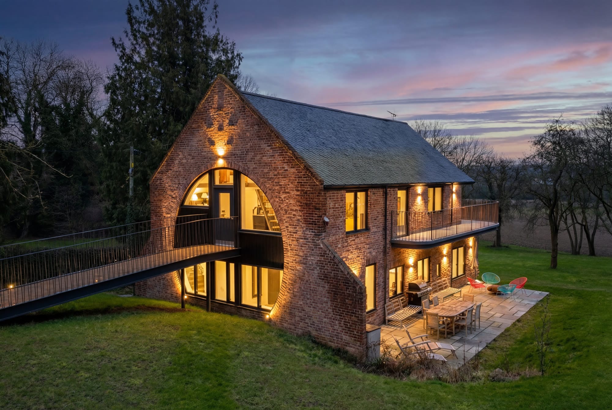 The Hollow, a brick barn house at twilight with an arched glass gable, a footbridge and a lit terrace